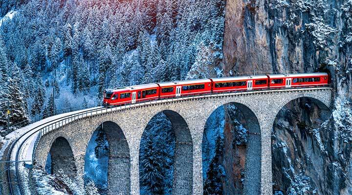 A red tourist train coming out of a mountain tunnel heading over a famous birdge in Europe.