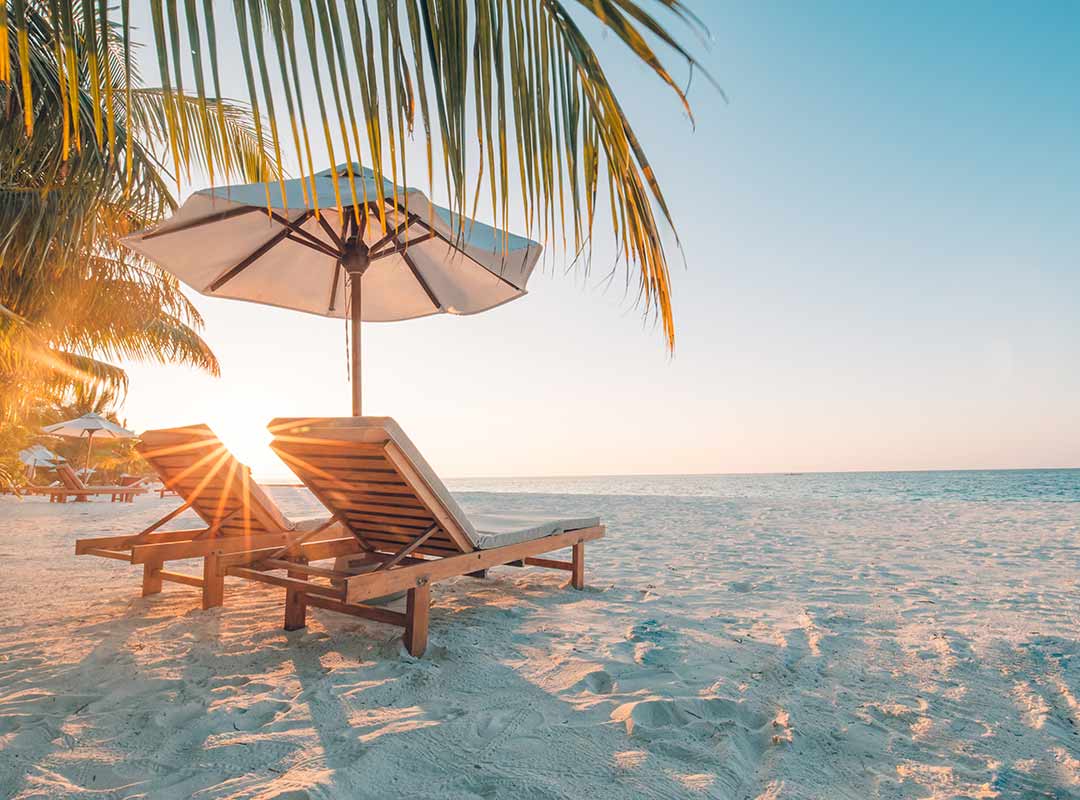 Two deckchairs on a white sand beach under a palm tree with the sun setting over the sea.