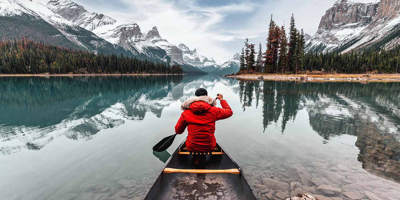 A man in a red jacket canoeing through a lake in canada on an adventure holiday, heading for the snowy mountains.