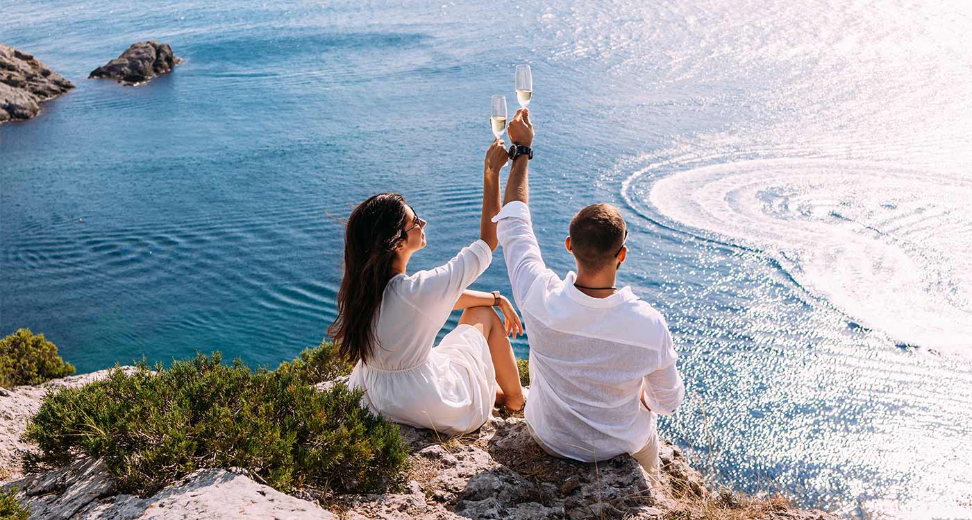 A couple celebrating their wedding anniversary with champagne by the sea, sitting on a rocky outcrop on a tropical island.
