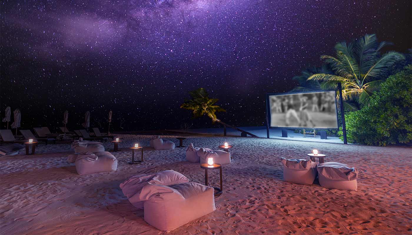 A moonlit cinema on a sandy beach by the sea, with bean bags, candles and palm trees.