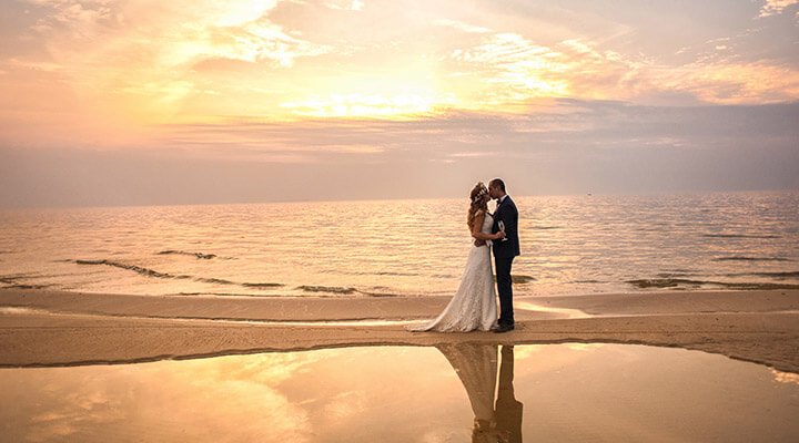 A couple exchanging vows barefoot on the beach with the sun setting over the waves.