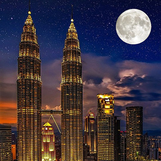 The petronas towers in kuala lumpur, malaysia, lit up at night beside a full moon.