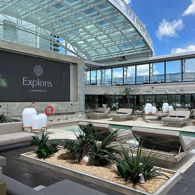 The large explora journey sign over the beautiful pool on the deck of the ship with sun loungers and palm trees dotted about.