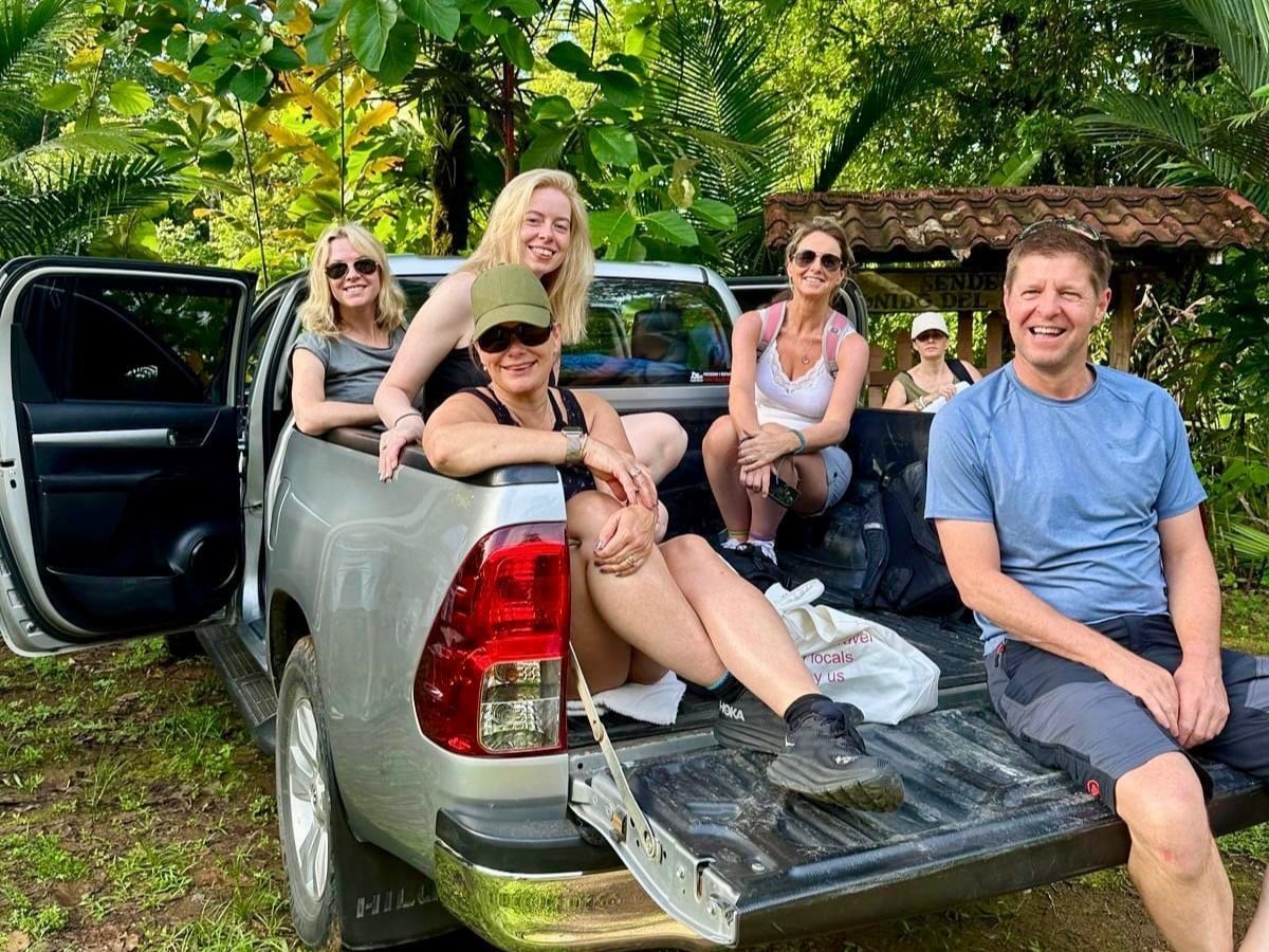 Group of happy travellers sitting on a pickup truck surrounded by lush tropical vegetation.