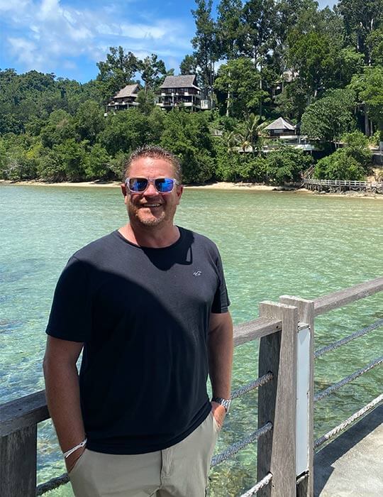 Mark Francis, Once in a Lifetime Holidays travel agent, standing by beautiful water and a villa.