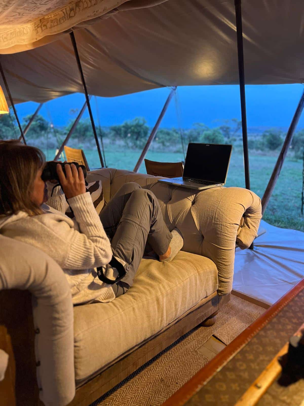 Person using binoculars in luxury safari tent with laptop nearby