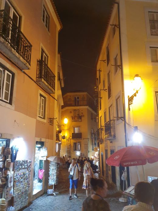 Narrow cobblestone street in Lisbon's Alfama district at night, illuminated by traditional lanterns with tourists browsing local shops