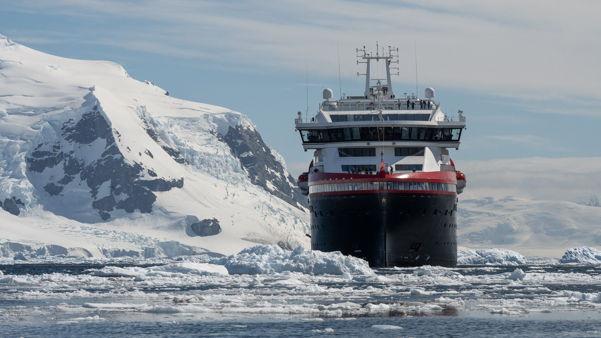 Expedition cruise ship navigating through ice flows in Antarctica with snow-covered mountains in background