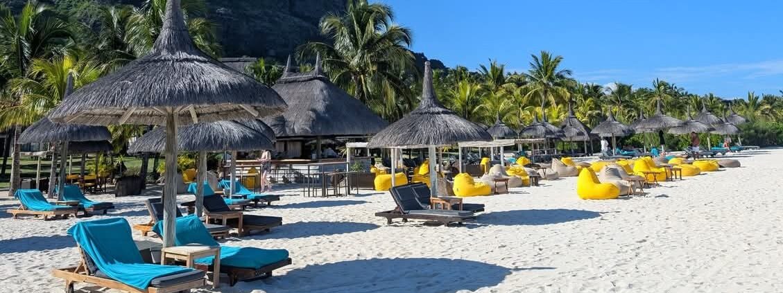 Pristine white sandy beach in Mauritius with thatched umbrellas, blue and yellow loungers, palm trees, and dramatic mountain backdrop.