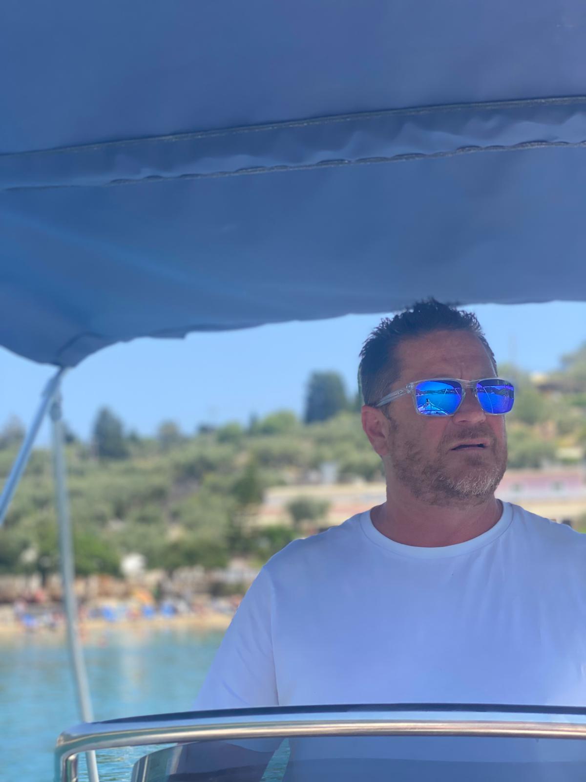Man in blue sunglasses and white shirt on boat with Mediterranean coastline and beach in background
