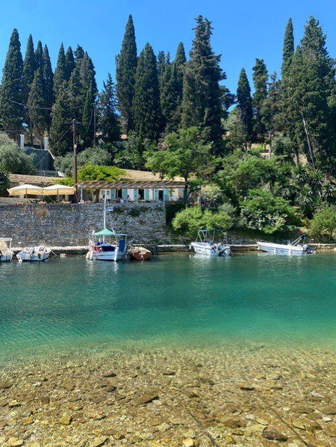 Crystal clear emerald waters of small Greek harbor with moored boats, stone quay, and cypress trees