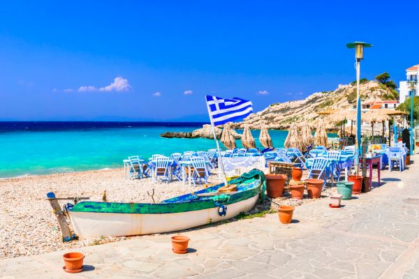 Greek beach taverna with traditional fishing boat, blue tables, and Greek flag overlooking turquoise sea