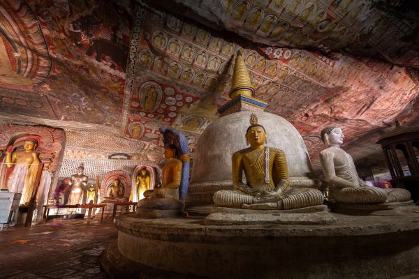 Interior of ancient Buddhist cave temple with golden Buddha statues, white stupa, and colorful ceiling frescoes