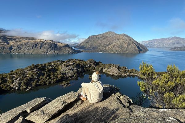 Person sitting on rocky outcrop overlooking vast mountain lake with barren hills and blue sky