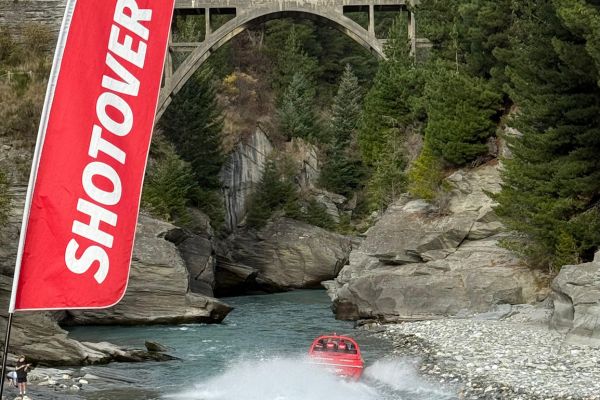 Red jet boat speeding through rocky river gorge creating water spray with bridge and canyon walls