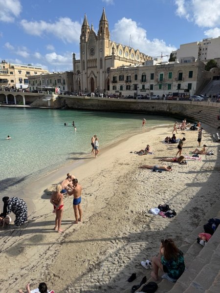 Turquoise Mediterranean waters and sandy beach in Malta with neo-gothic church overlooking swimmers and sunbathers enjoying Malta beaches on sunny summer day