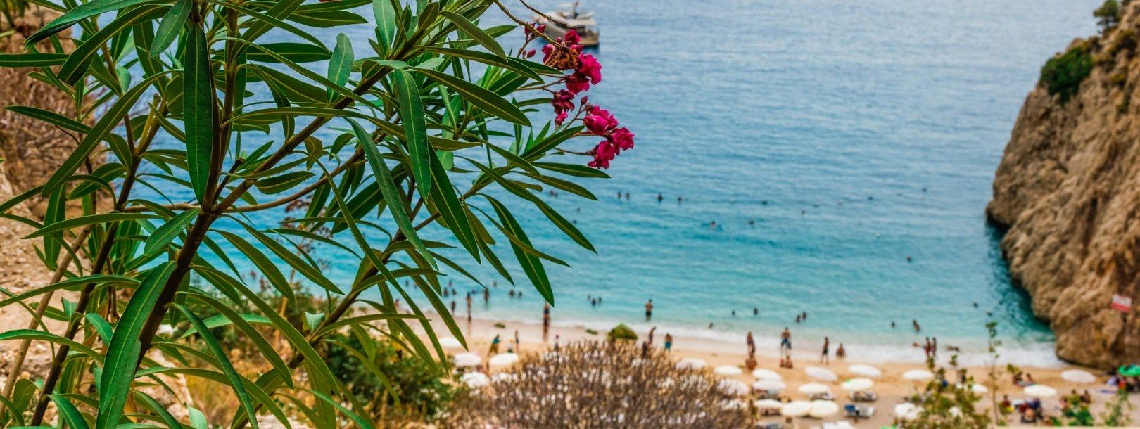 Turquoise Mediterranean waters and sandy beach in Kalkan Turkey with pink bougainvillea flowers, dramatic cliffs and swimmers enjoying Turquoise Coast luxury holiday destination