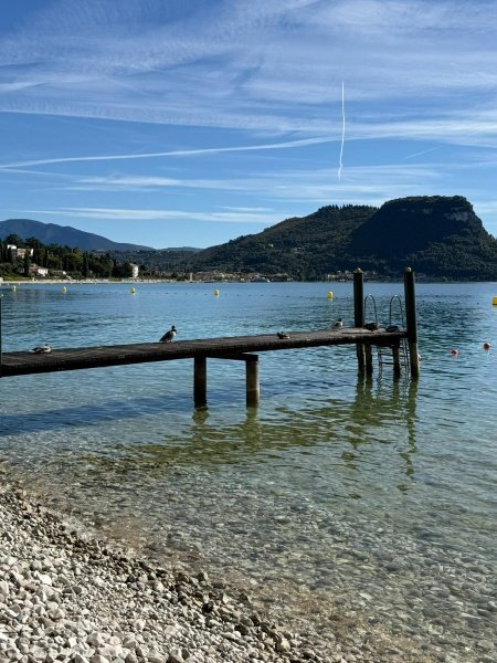 Wooden pier on Lake Garda with crystal-clear turquoise waters, pebble beach and mountain backdrop showcasing peaceful Italian lake escape and luxury slow travel destination