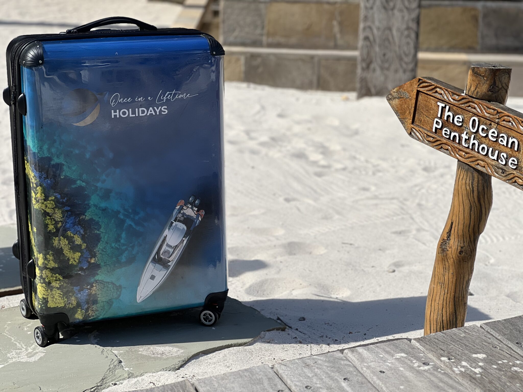 Once in a Lifetime Holidays branded suitcase on a sandy beach beside an Ocean Penthouse sign