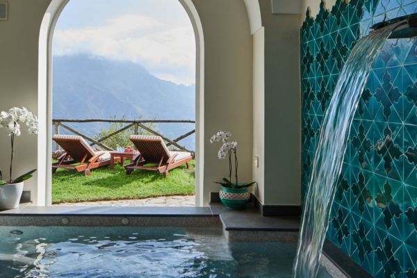 Indoor spa pool with teal mosaic waterfall and arched window overlooking Amalfi Coast mountains at Palazzo Avino